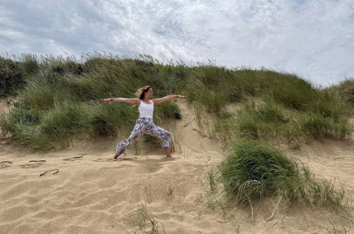Frau praktiziert die Yoga-Standhaltung Krieger II (Virabhadrasana II) barfuß am Strand vor einer Dünenlandschaft unter leicht bewölktem Himmel.