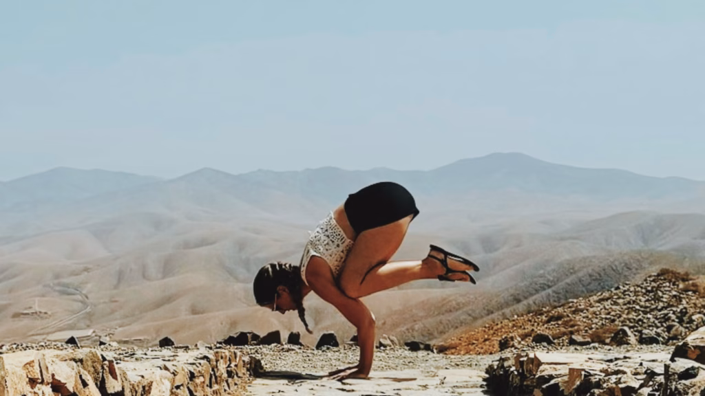 Sarah Frößl übt eine Krähe-Pose (Bakasana) auf einem steinigen Plateau vor einer weitläufigen Wüsten- und Berglandschaft unter klarem Himmel.