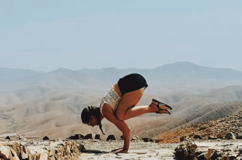 Sarah Frößl übt eine Krähe-Pose (Bakasana) auf einem steinigen Plateau vor einer weitläufigen Wüsten- und Berglandschaft unter klarem Himmel.